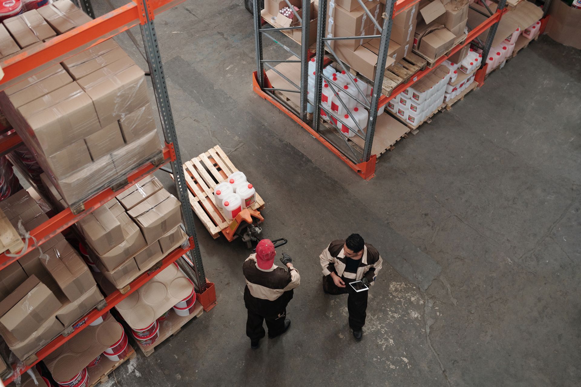 Stock photograph of two warehouse workers beside a pallet jack and storage racks